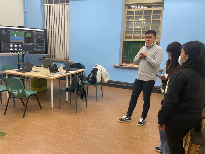 A team member presenting the project in a classroom, audience listening, the Unity editor visible on a monitor in the background.