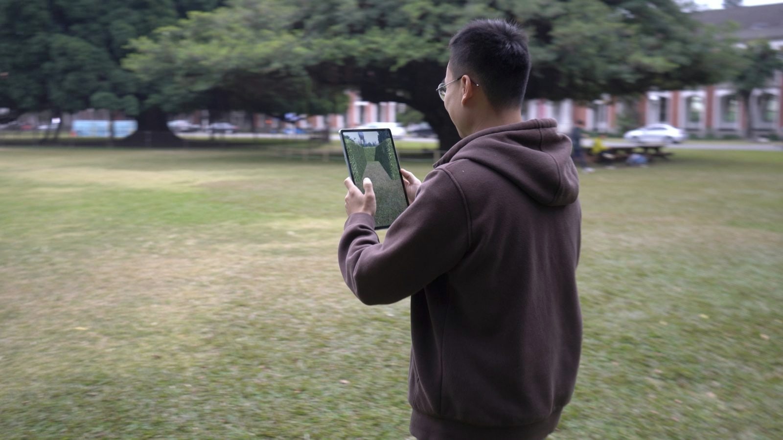 A tester from behind, holding a tablet that shows the AR maze; he stands on the real grass with banyan in the background.