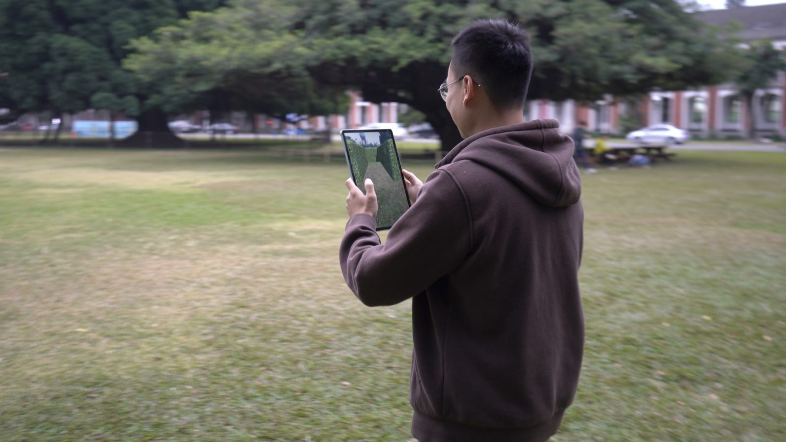 A tester from behind, holding a tablet that shows the AR maze; he stands on the real grass with banyan in the background.