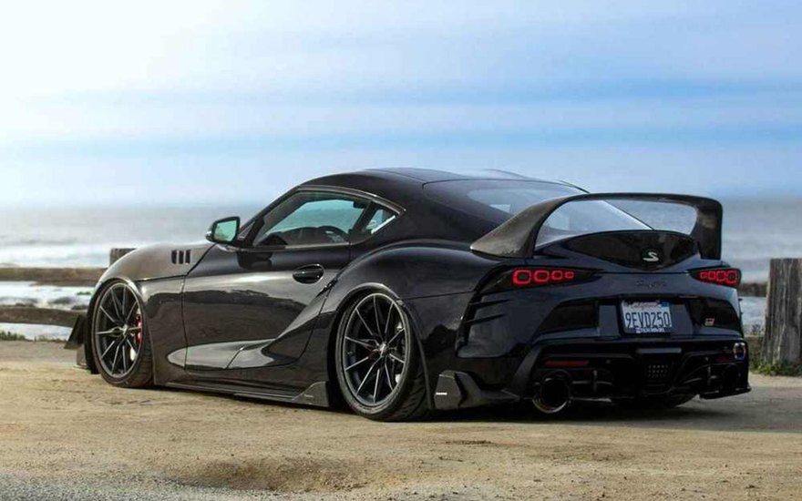 A black Toyota Supra with ARMASPEED carbon fiber kit parked at a coastal road, rear three-quarter view.