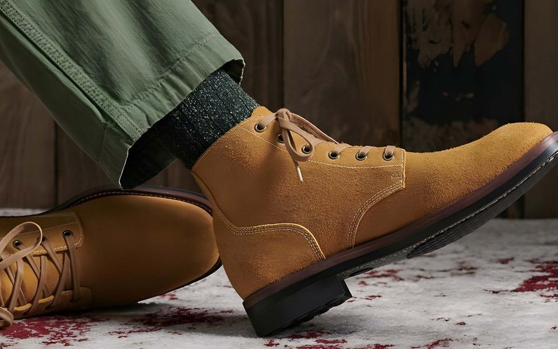 A tan suede chukka boot on green chinos against a dark wood-plank backdrop — brand footwear line.
