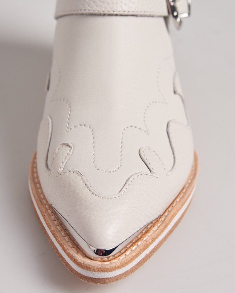 Buckle Boots: a single white pebbled-leather Western ankle boot in side profile on a clean white studio backdrop, showing the twin silver buckle straps, the cut-out chelsea panel, the metal toe cap, and the stacked wood heel.