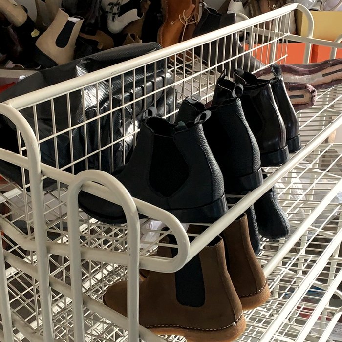 Finished black chelsea boots arranged on a white wire shelving rack with more boot stock visible on background shelves — full-size samples staged for review.