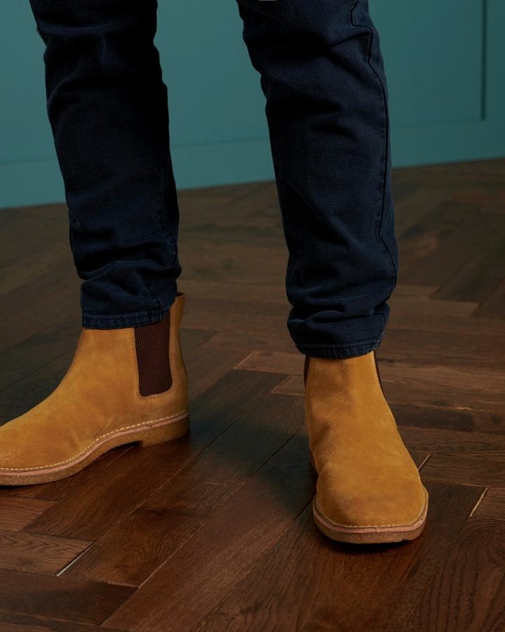 Mustard-suede Chelsea ankle boot on dark herringbone parquet, paired with dark navy denim.