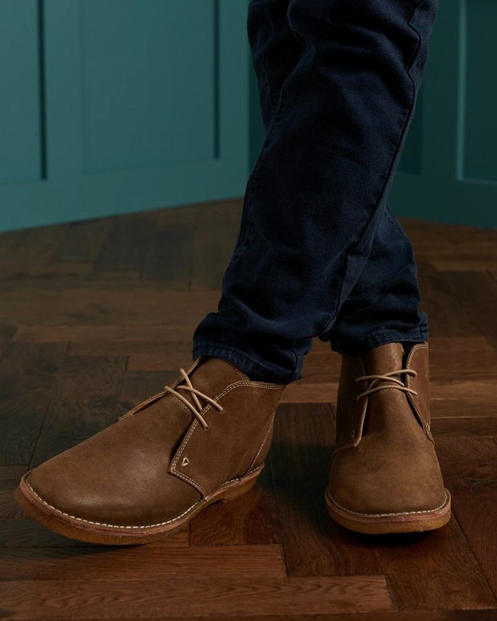 Brown suede chukka boot on dark herringbone parquet, paired with dark navy trousers.