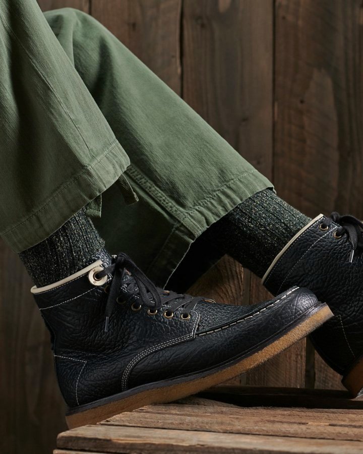 Black pebbled-leather work boot with cream eyelet ring, on green chinos and wood plank.