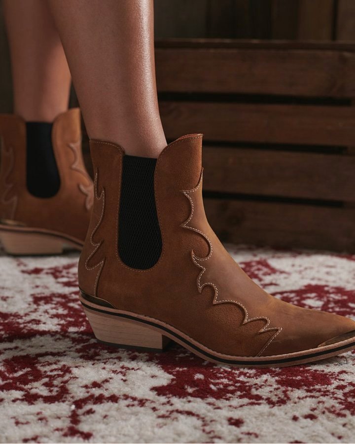 Tan suede Western Chelsea boot with stitched silhouette and contrasting heel cup, on red rug.