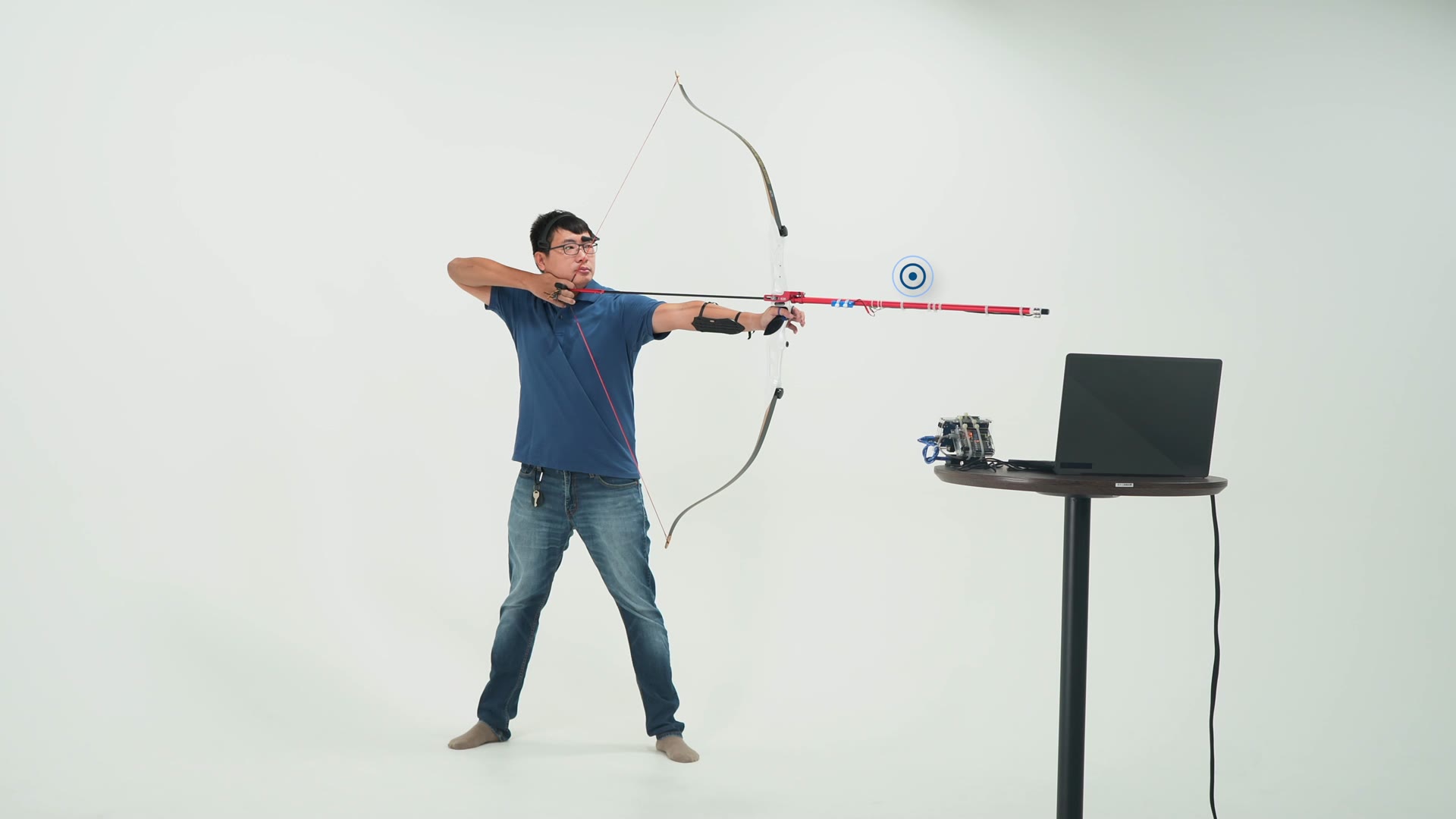 A participant draws a modified recurve bow in a white studio; a laptop on a stand beside him shows the live feedback.